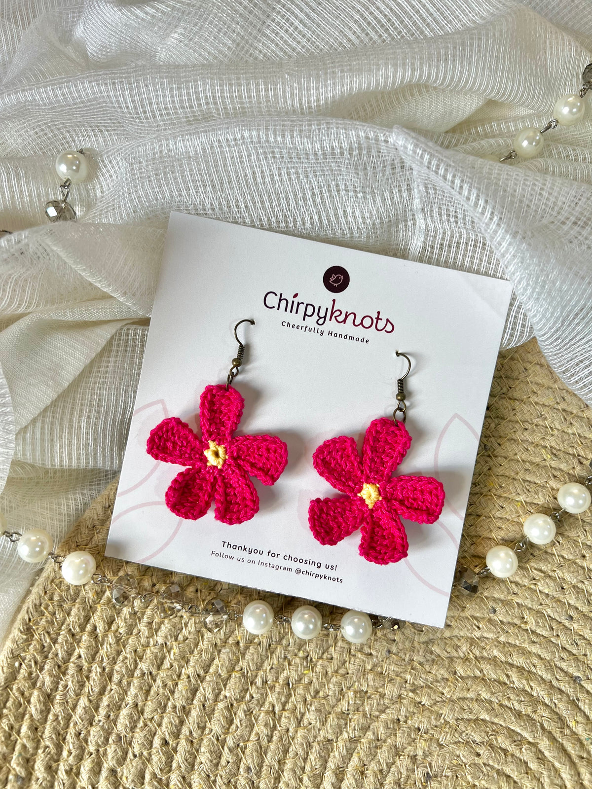 A pair of berry pink crocheted, 5 petal flower-shaped dangle earrings by Chirpyknots, displayed on a white brand card over a woven mat and white fabric and pearl beads.