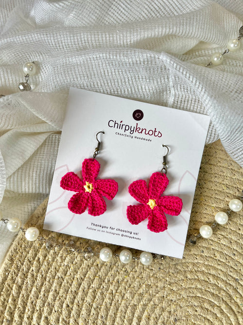 A pair of berry pink crocheted, 5 petal flower-shaped dangle earrings by Chirpyknots, displayed on a white brand card over a woven mat and white fabric and pearl beads.