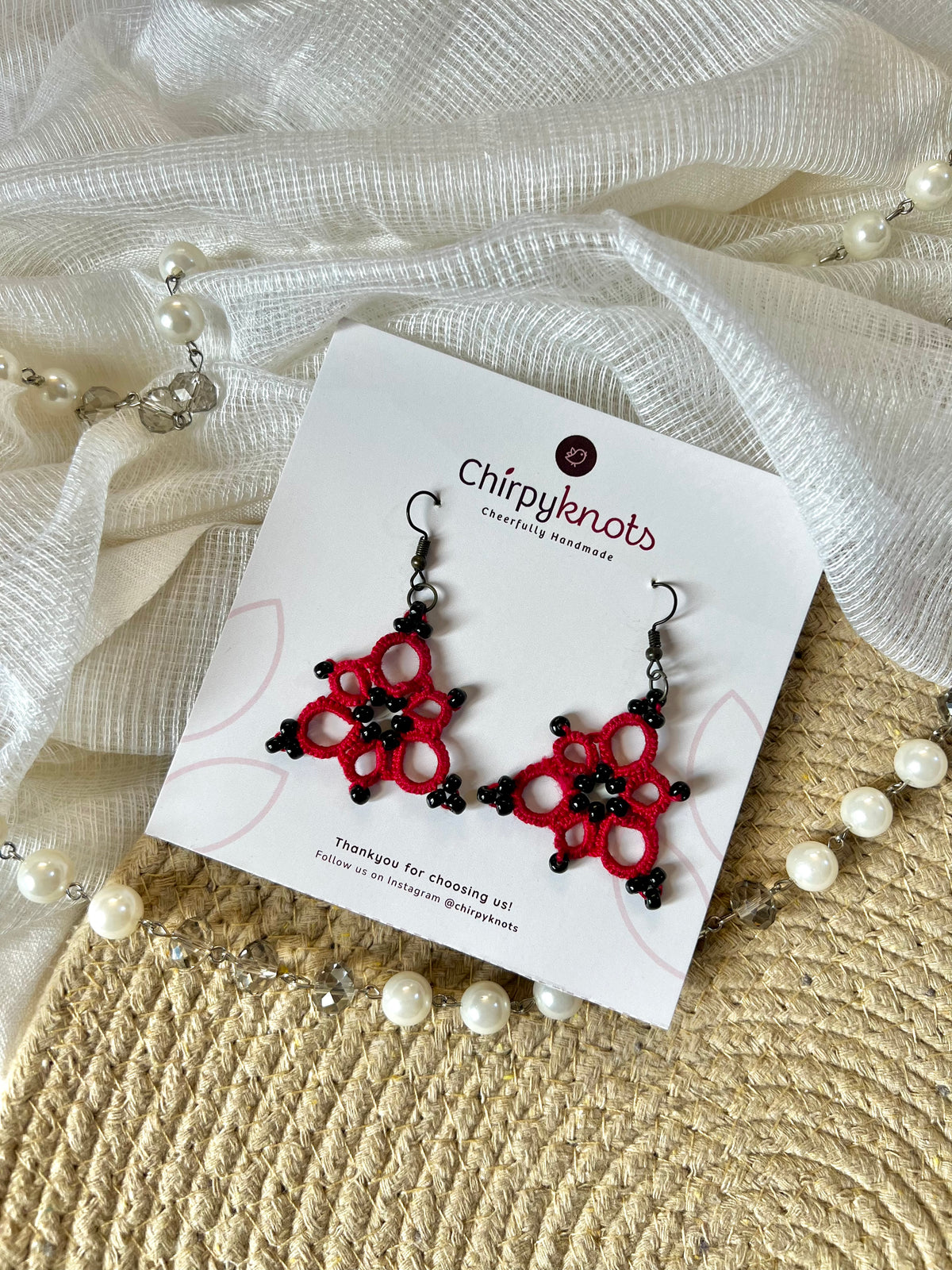 A pair of maroon and black tatted, triangle-shaped beaded dangle earrings by Chirpyknots, displayed on a white brand card over a woven mat and white fabric and pearl beads.