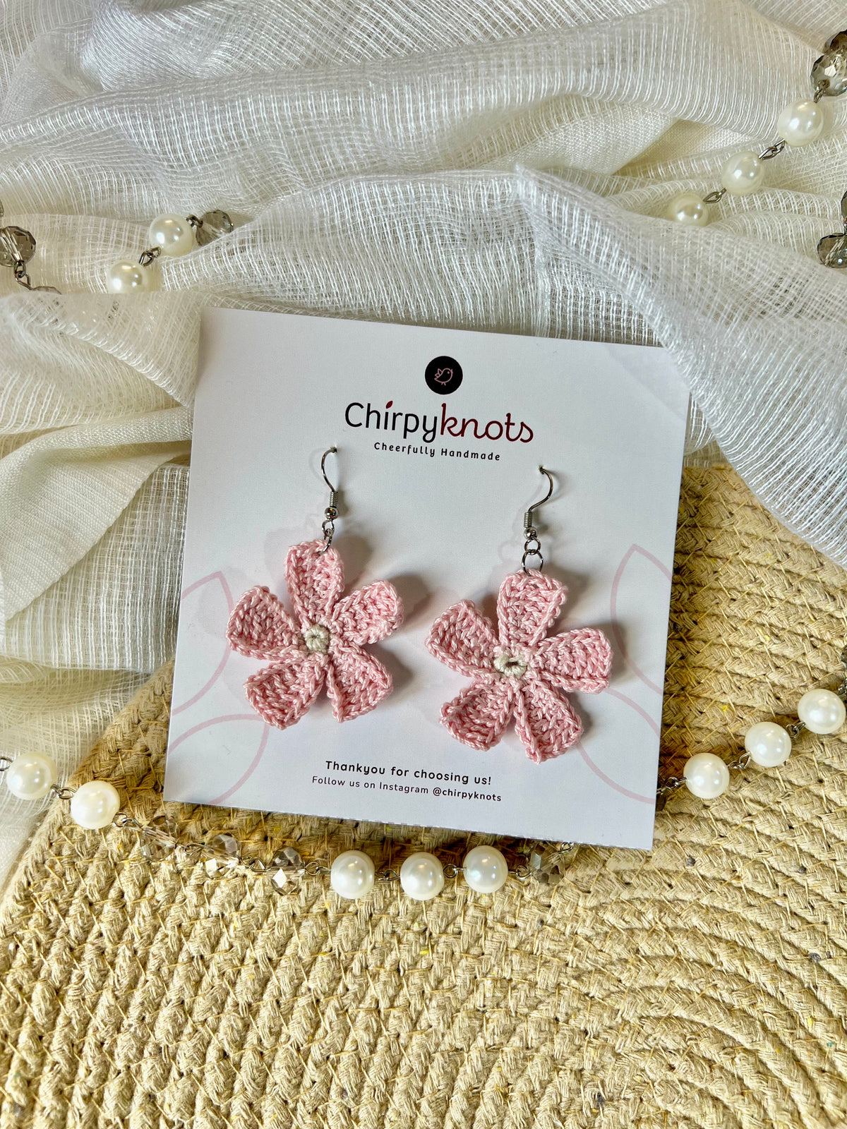 A pair of light pink crocheted, 5 petal flower-shaped dangle earrings by Chirpyknots, displayed on a white brand card over a woven mat and white fabric and pearl beads.