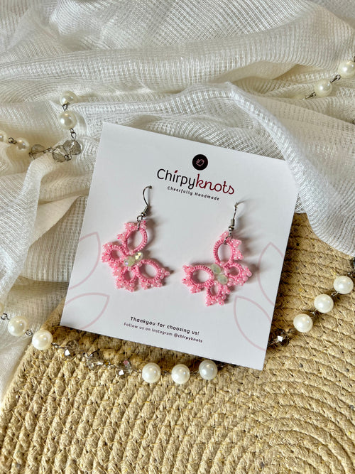 A pair of tatted pink, beaded, butterfly style dangle earrings by Chirpyknots, displayed on a white brand card over a woven mat and white fabric and pearl beads.