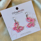 A closeup of a pair of tatted pink, beaded, butterfly style dangle earrings by Chirpyknots, displayed on a white brand card.