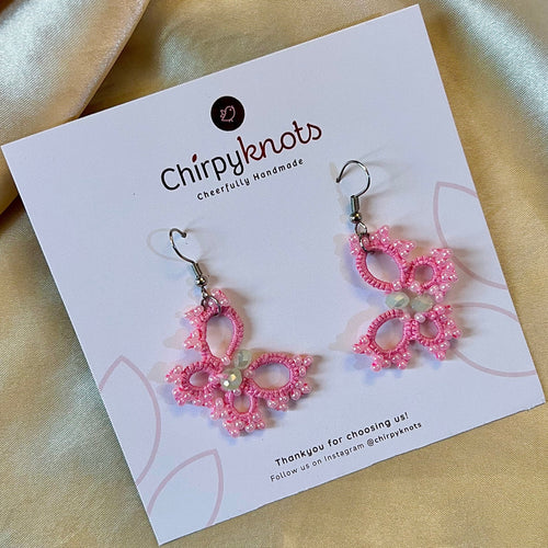 A closeup of a pair of tatted pink, beaded, butterfly style dangle earrings by Chirpyknots, displayed on a white brand card.