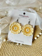 A pair of yellow and cream crocheted, 12 petal daisy flower-shaped round, dangle earrings by Chirpyknots, displayed on a white brand card over a woven mat and white fabric and pearl beads.