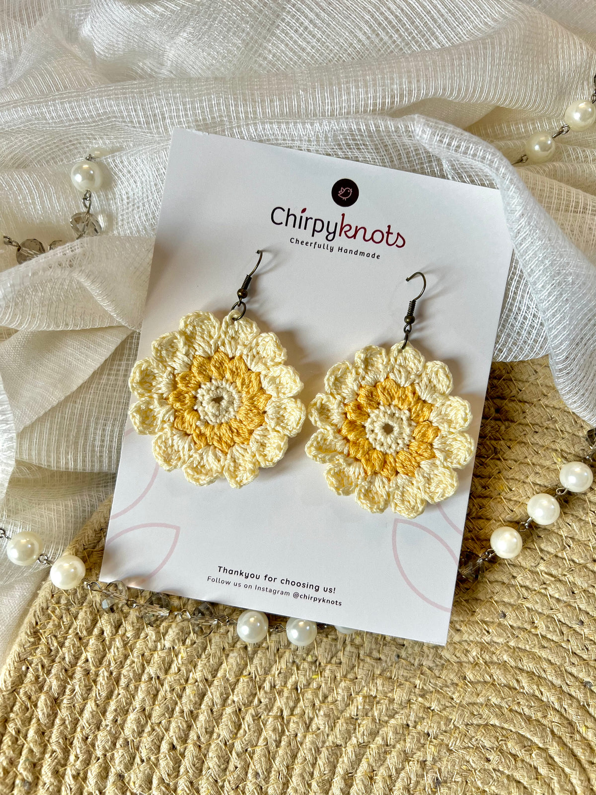 A pair of yellow and cream crocheted, 12 petal daisy flower-shaped round, dangle earrings by Chirpyknots, displayed on a white brand card over a woven mat and white fabric and pearl beads.