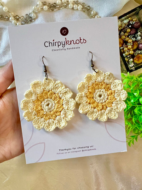 A closeup of a pair of yellow and cream crocheted, 12 petal daisy flower-shaped round, dangle earrings by Chirpyknots, displayed on a white brand card.