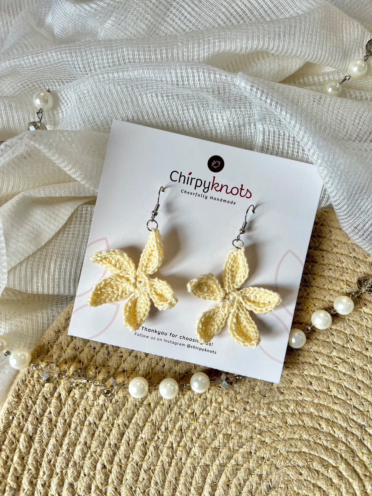 A pair of butter yellow crocheted, flower-shaped dangle earrings by Chirpyknots, displayed on a white brand card over a woven mat and white fabric and pearl beads.