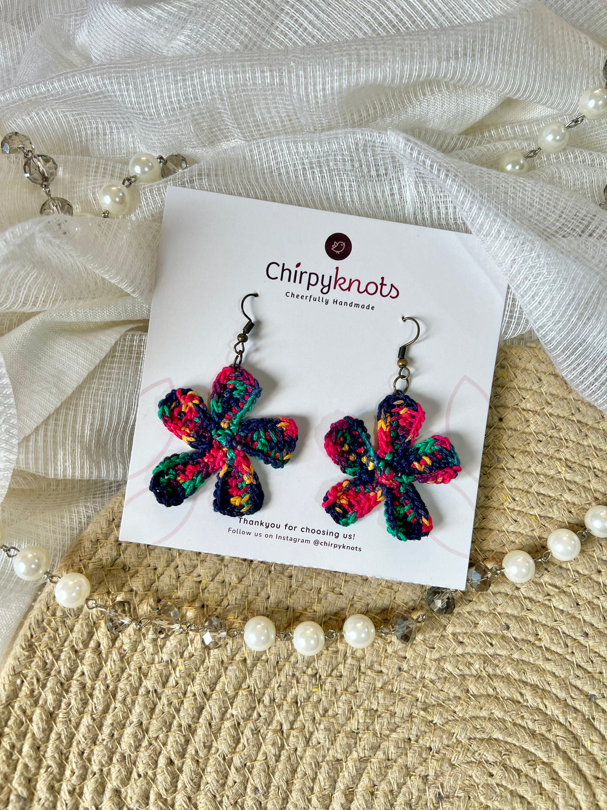 A pair of dark multicolor crocheted, 5 petal flower-shaped dangle earrings by Chirpyknots, displayed on a white brand card over a woven mat and white fabric and pearl beads.