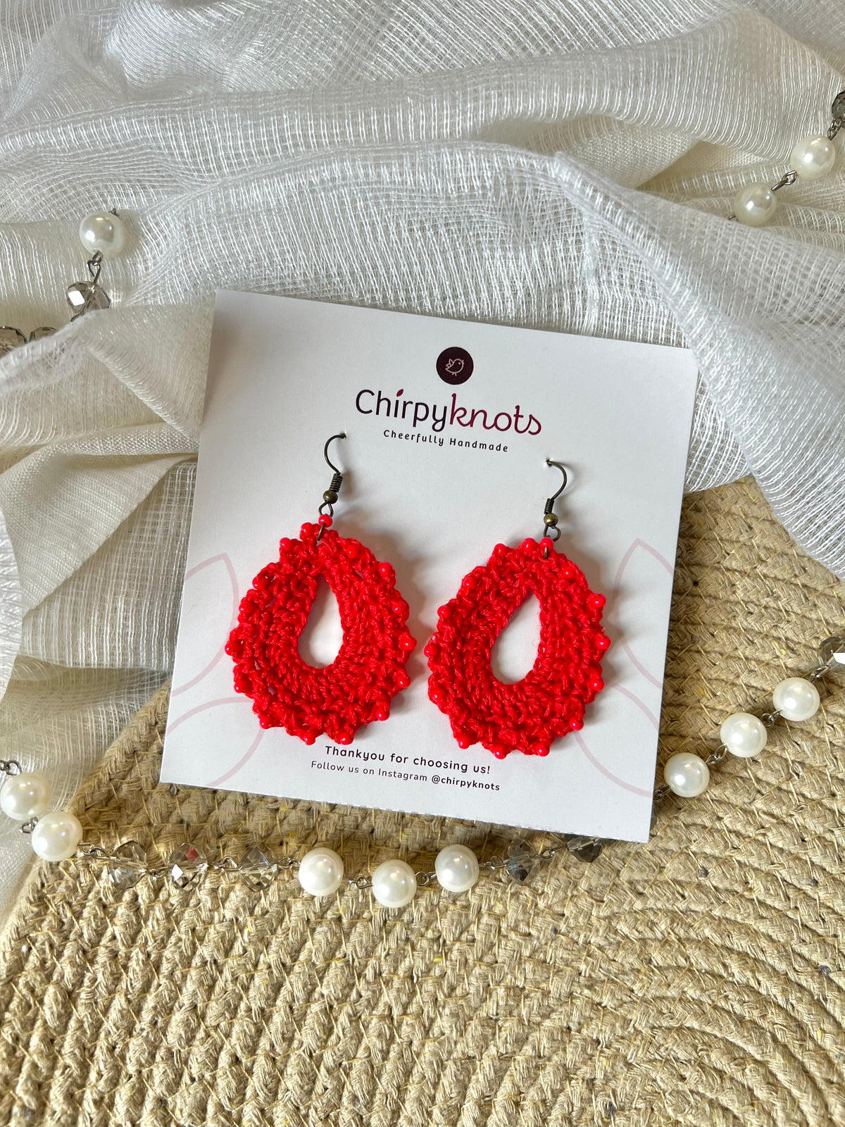 A pair of crocheted red, oval shaped beaded dangle earrings by Chirpyknots, displayed on a white brand card over a woven mat and white fabric and pearl beads.