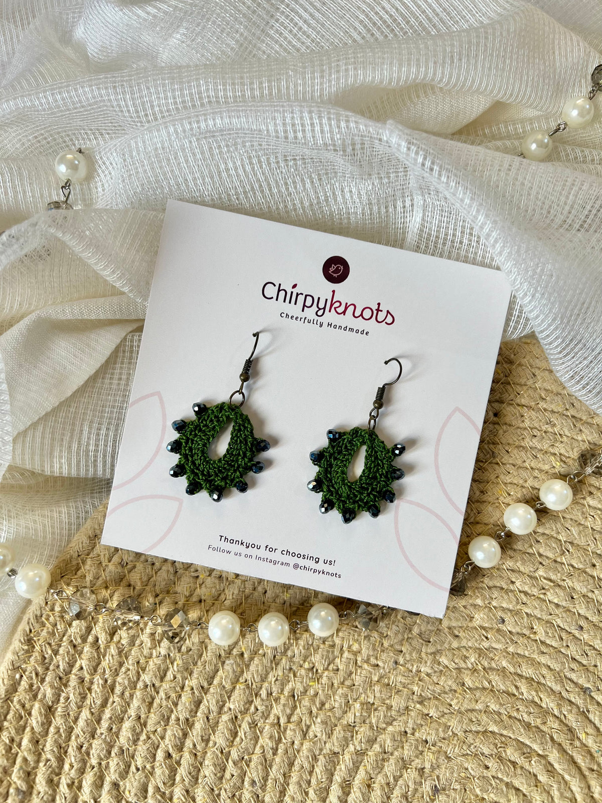 A pair of crocheted dark green, small, oval shaped beaded dangle earrings by Chirpyknots, displayed on a white brand card over a woven mat and white fabric and pearl beads.