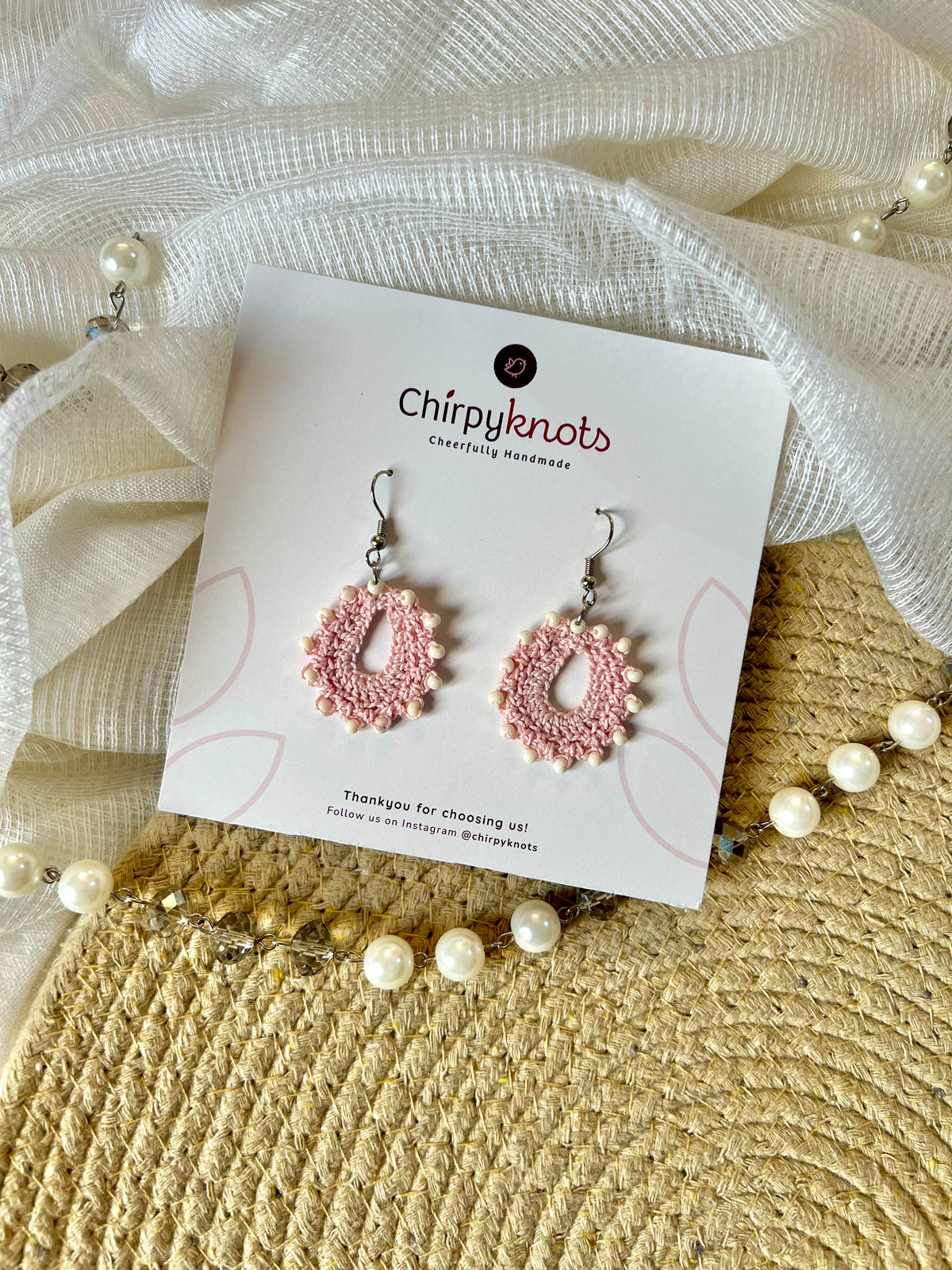 A pair of crocheted light pink, small, oval shaped beaded dangle earrings by Chirpyknots, displayed on a white brand card over a woven mat and white fabric and pearl beads.