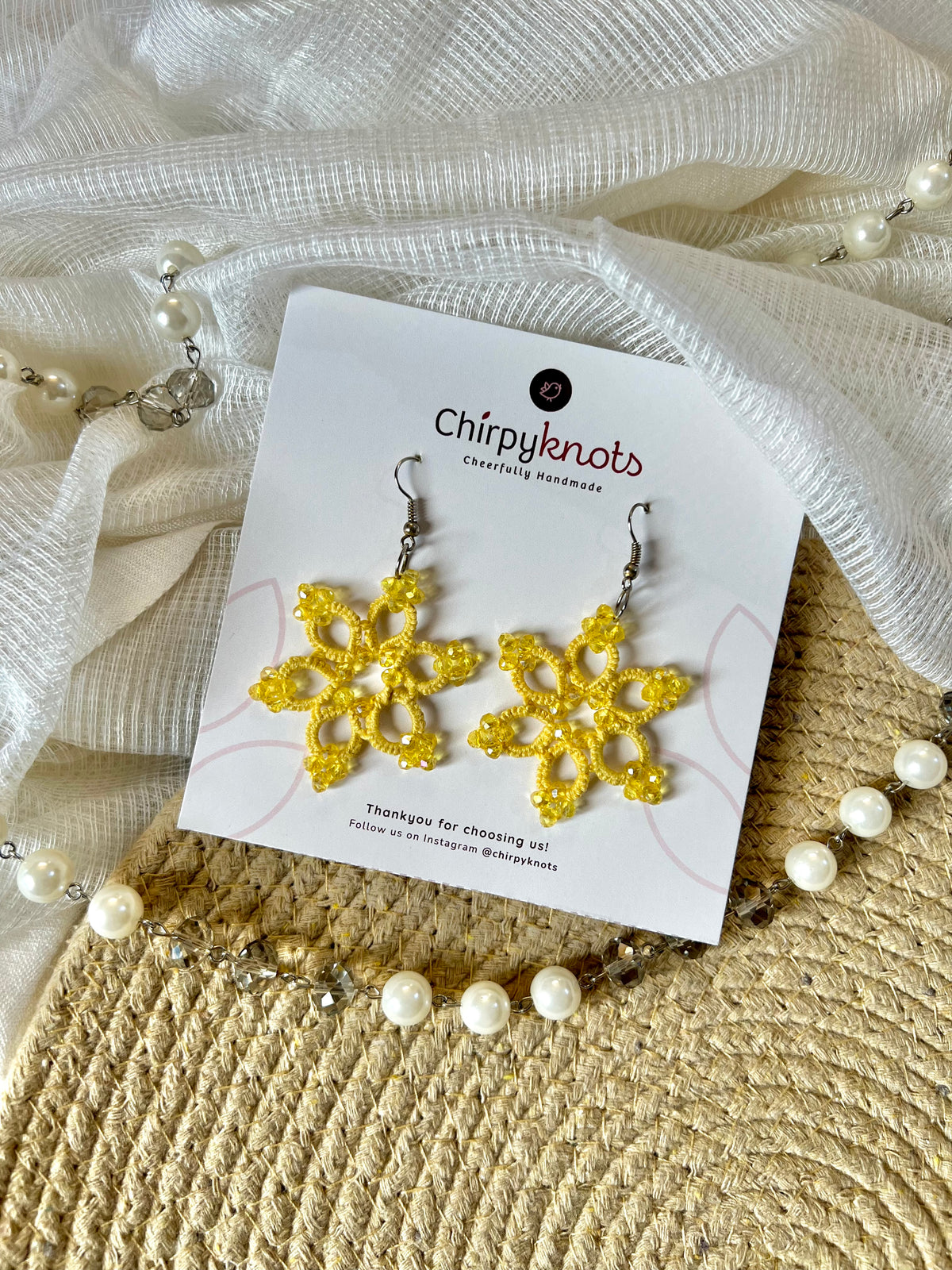A pair of yellow tatted, 6 petal star flower shaped beaded dangle earrings by Chirpyknots, displayed on a white brand card over a woven mat and white fabric and pearl beads.