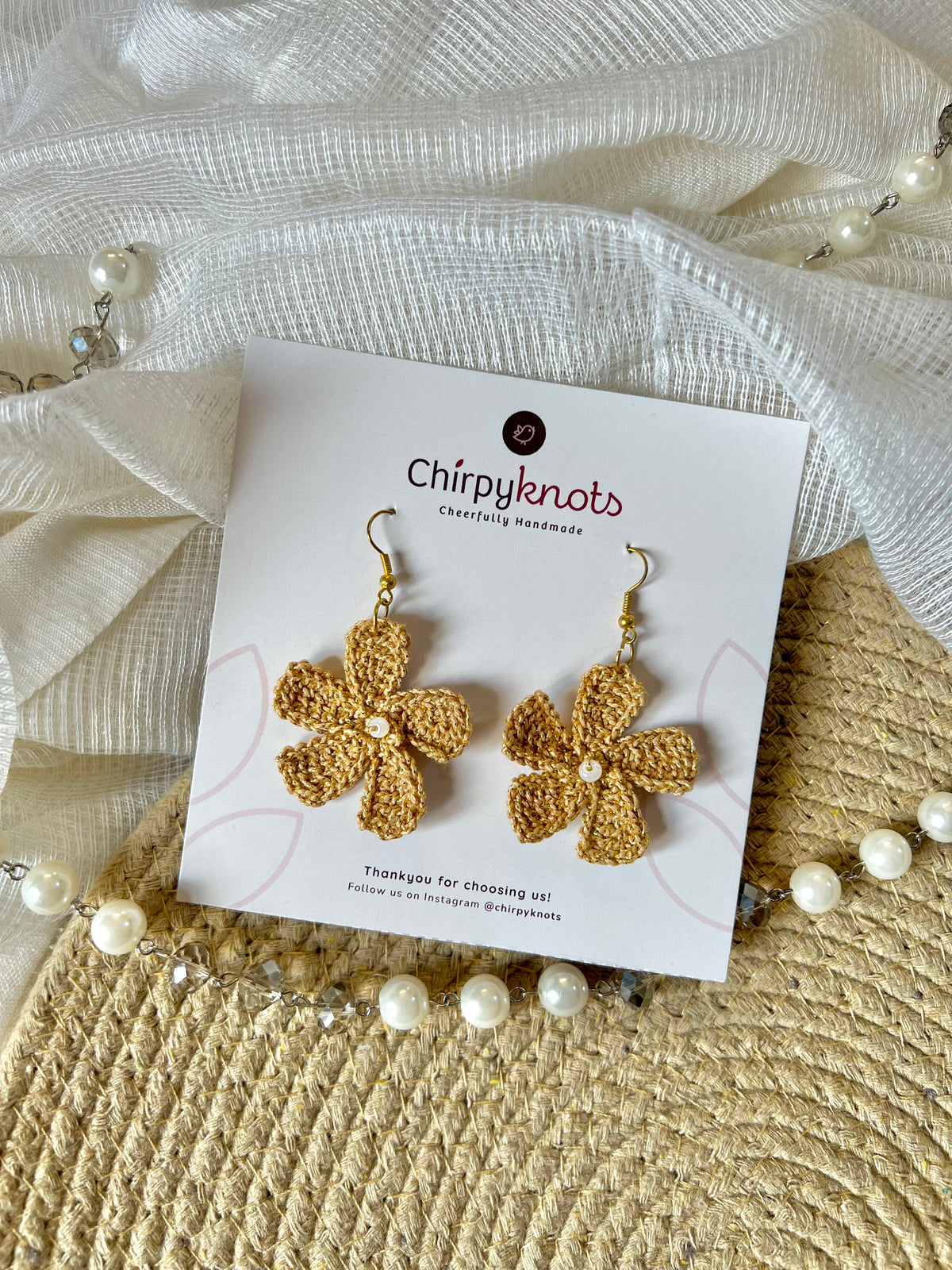 A pair of golden crocheted, 5 petal flower-shaped dangle earrings by Chirpyknots, displayed on a white brand card over a woven mat and white fabric and pearl beads.
