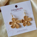 A closeup of a pair of golden crocheted, 5 petal flower-shaped dangle earrings by Chirpyknots, displayed on a white brand card.