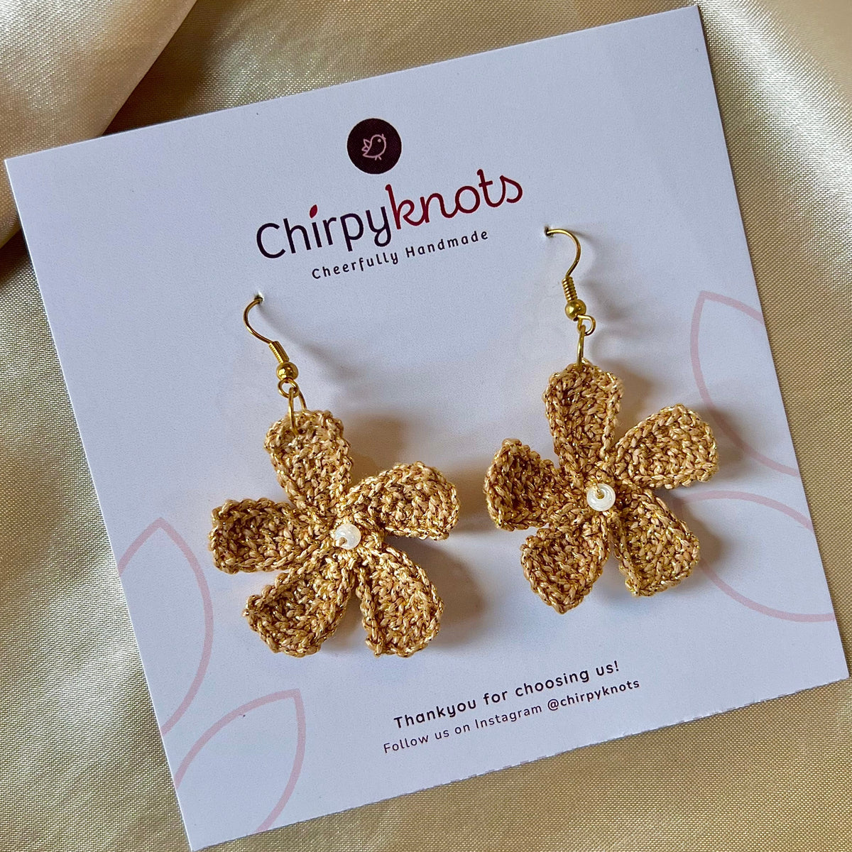 A closeup of a pair of golden crocheted, 5 petal flower-shaped dangle earrings by Chirpyknots, displayed on a white brand card.