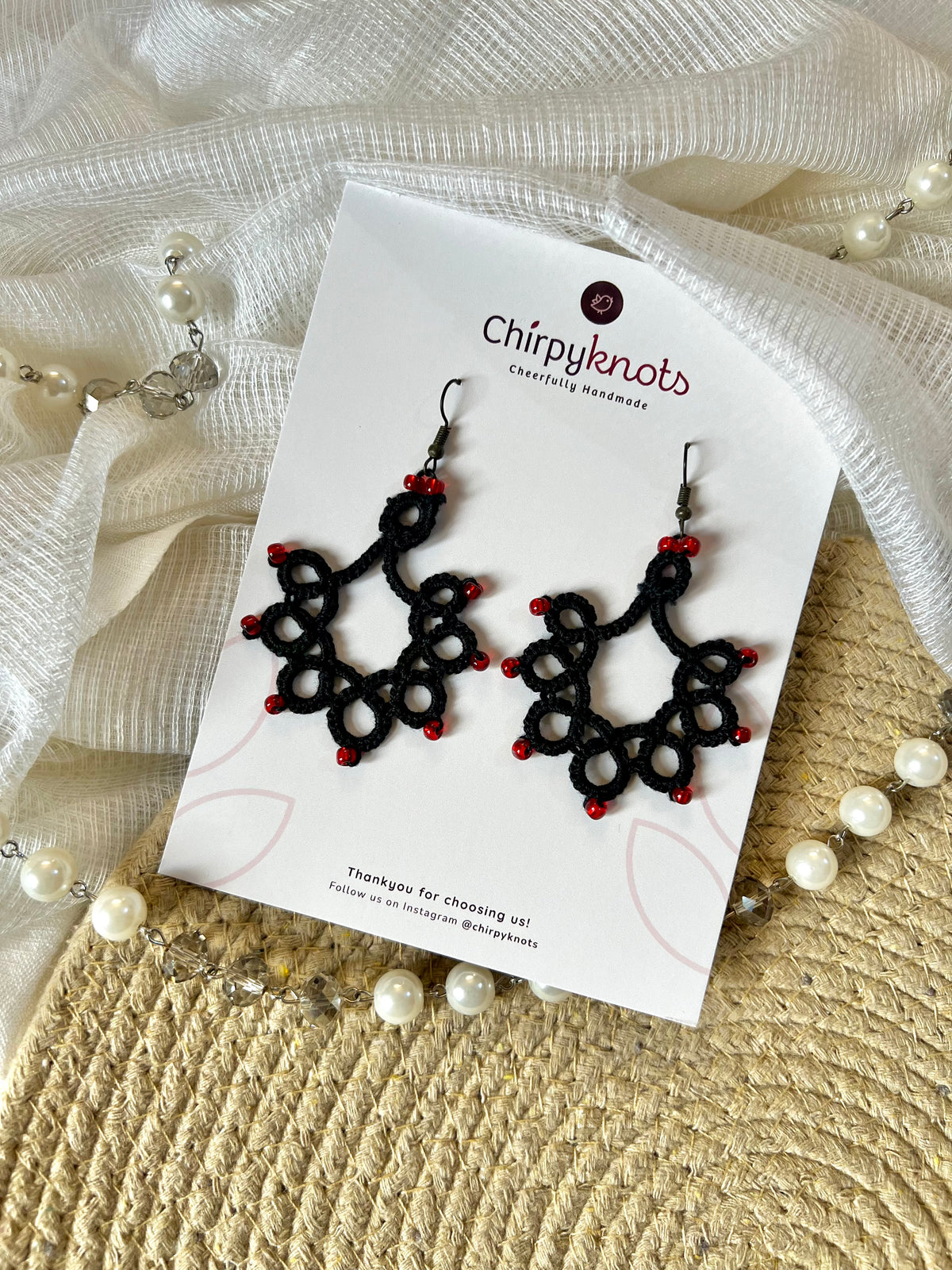 A pair of black tatted, chandelier beaded dangle earrings by Chirpyknots, displayed on a white brand card over a woven mat and white fabric and pearl beads.