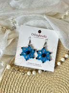 A pair of blue tatted, floral beaded dangle earrings by Chirpyknots, displayed on a white brand card over a woven mat and white fabric and pearl beads.