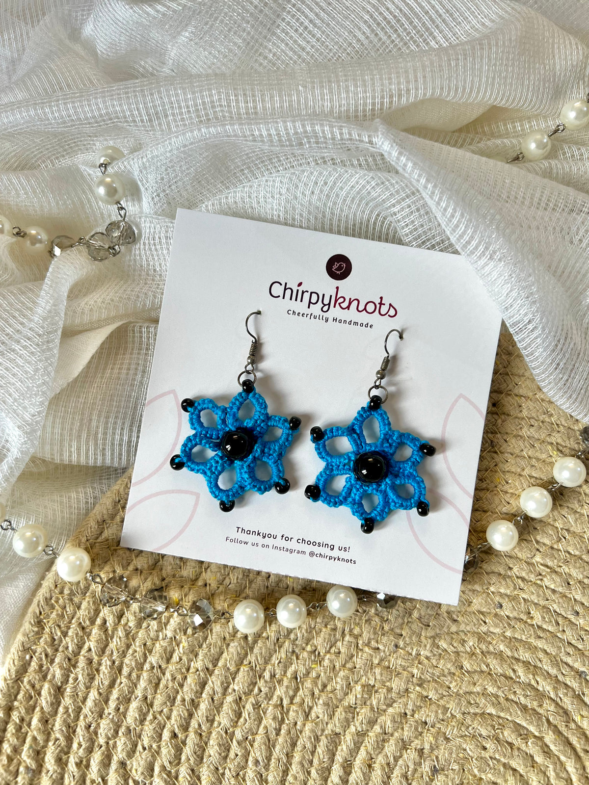 A pair of blue tatted, floral beaded dangle earrings by Chirpyknots, displayed on a white brand card over a woven mat and white fabric and pearl beads.