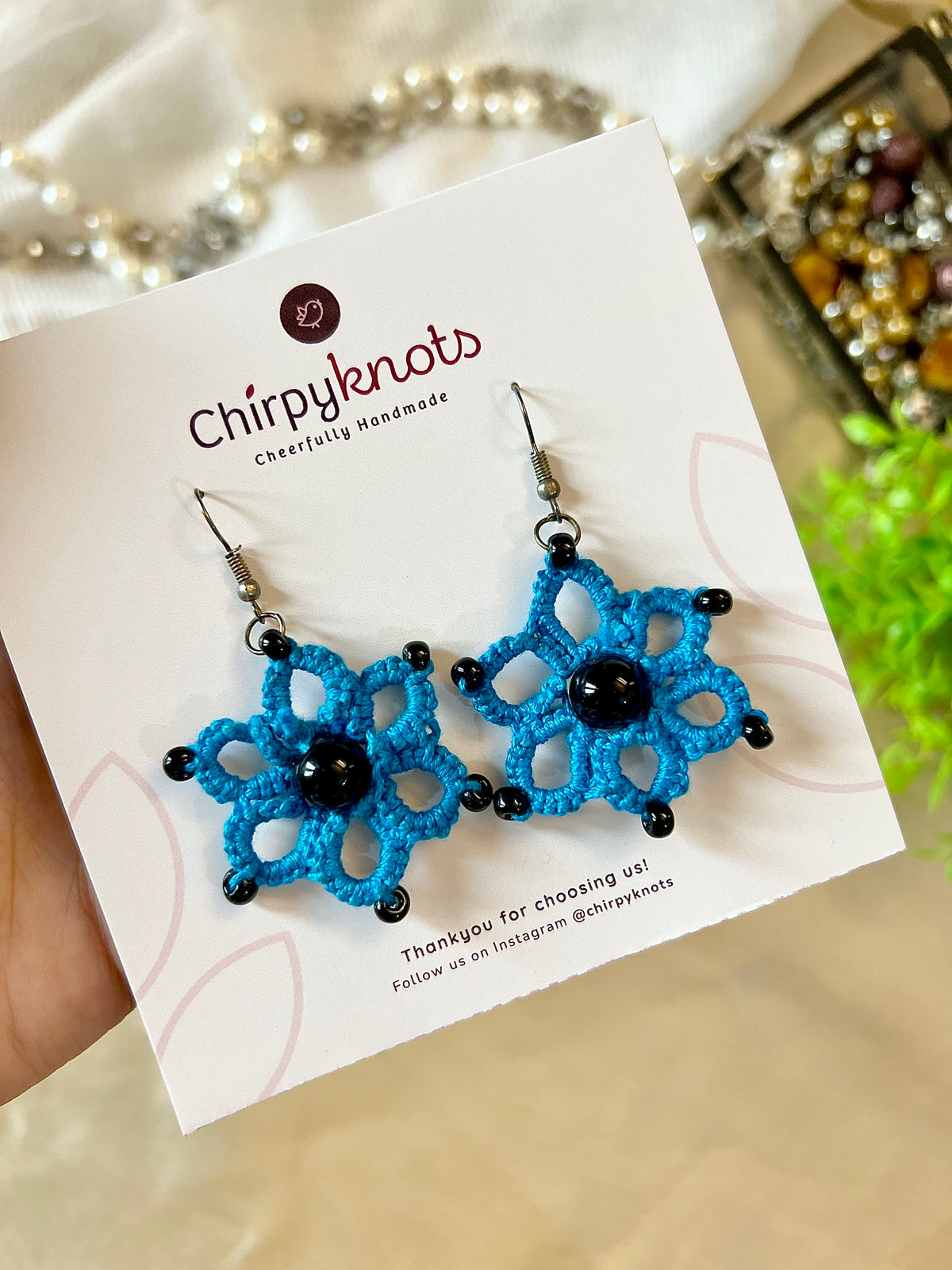 A closeup of a pair of blue tatted, floral beaded dangle earrings by Chirpyknots, displayed on a white brand card.