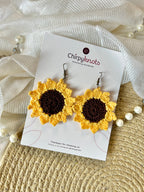 A pair of crocheted, sunflower dangle earrings by Chirpyknots, displayed on a white brand card over a woven mat and white fabric and pearl beads.