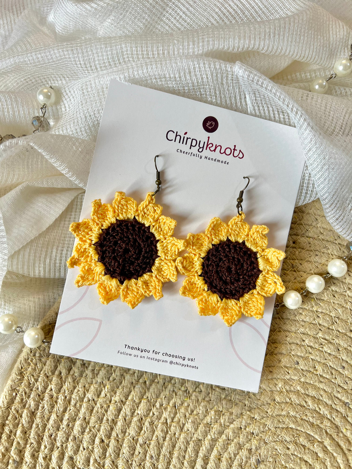 A pair of crocheted, sunflower dangle earrings by Chirpyknots, displayed on a white brand card over a woven mat and white fabric and pearl beads.