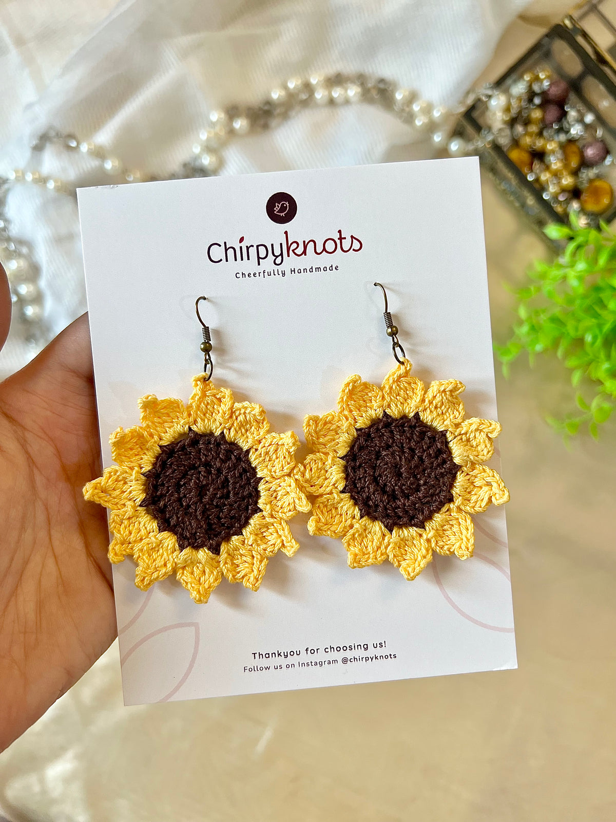 A closeup of a pair of crocheted, sunflower dangle earrings by Chirpyknots, displayed on a white brand card.
