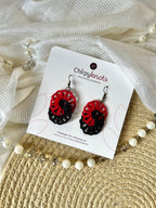 A pair of maroon and black tatted, infinity dangle earrings by Chirpyknots, displayed on a white brand card over a woven mat and white fabric and pearl beads.