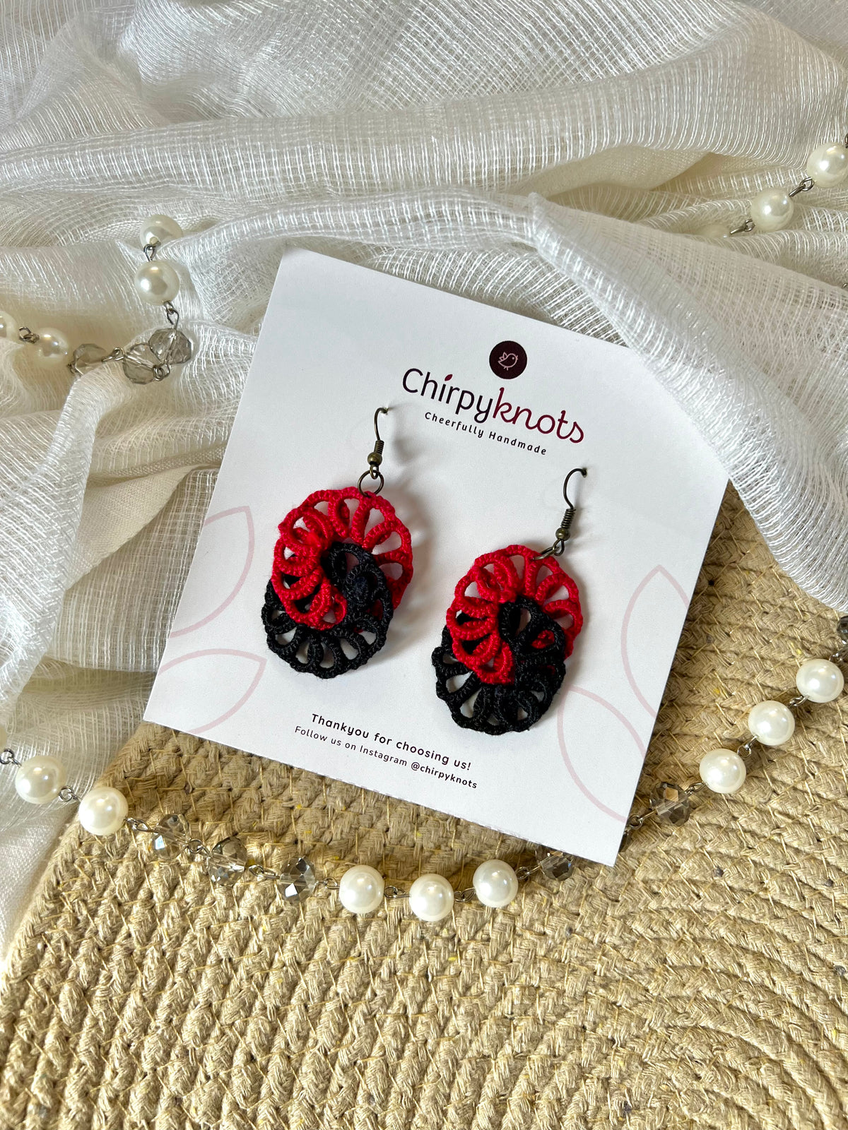 A pair of maroon and black tatted, infinity dangle earrings by Chirpyknots, displayed on a white brand card over a woven mat and white fabric and pearl beads.