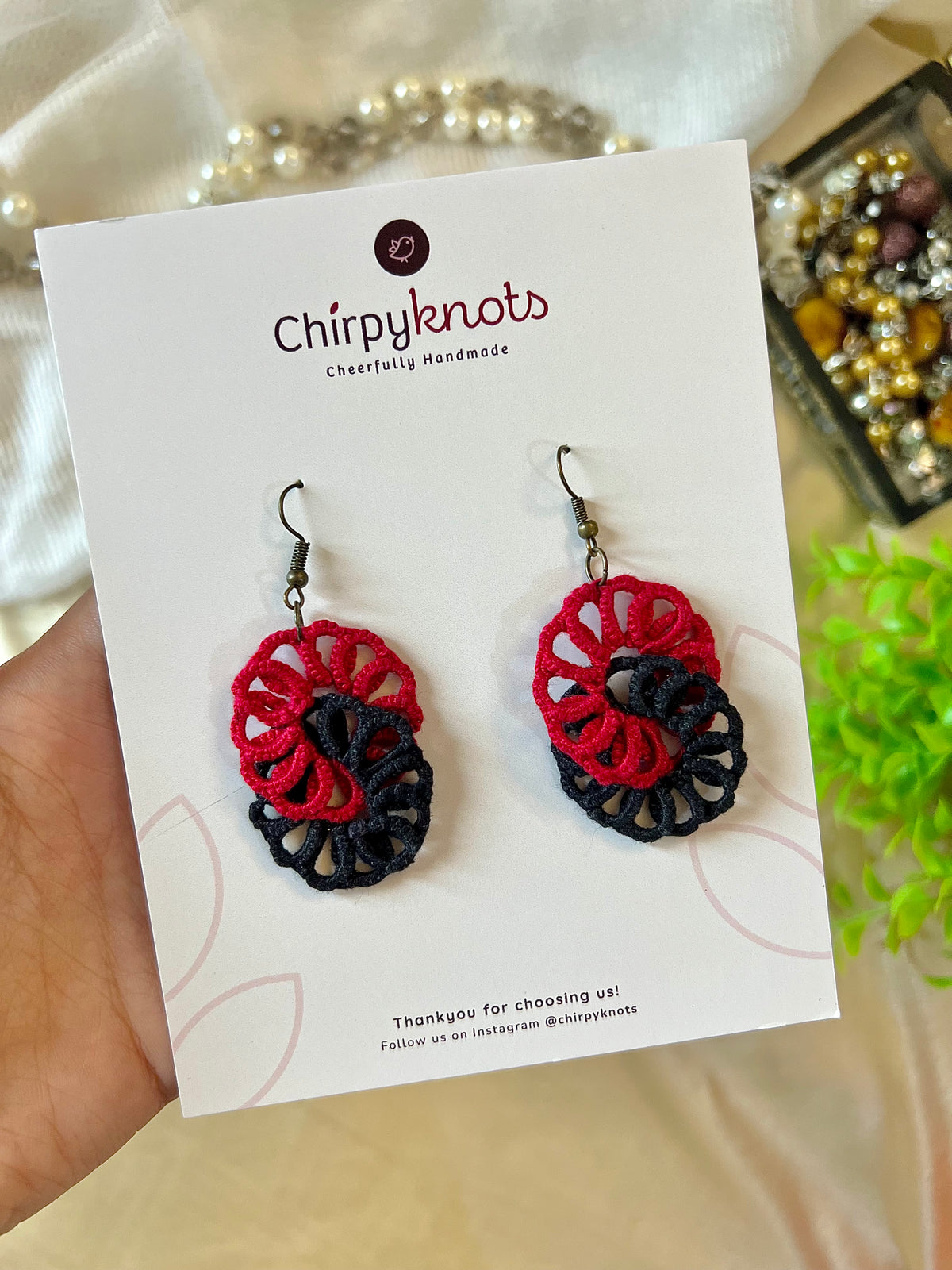 A closeup of a pair of maroon and black tatted, infinity dangle earrings by Chirpyknots, displayed on a white brand card.