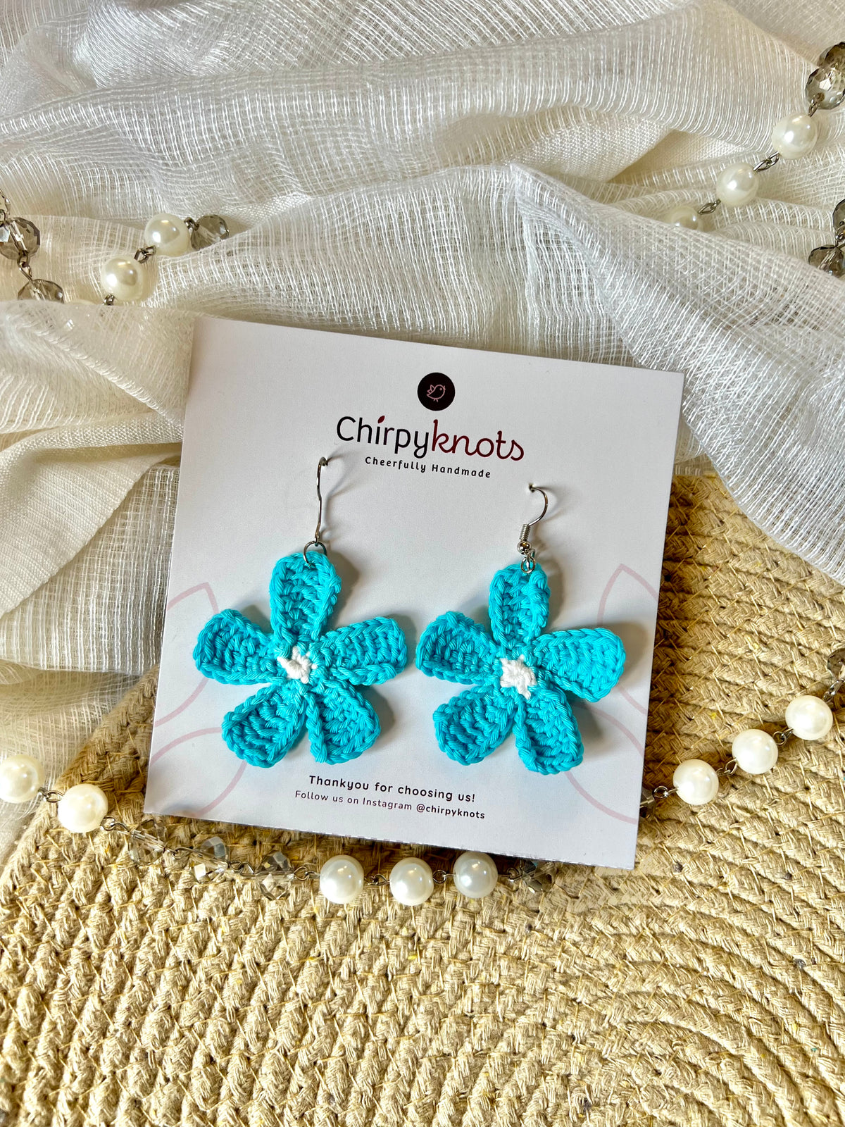 A pair of bright sea blue crocheted, 5 petal flower-shaped dangle earrings by Chirpyknots, displayed on a white brand card over a woven mat and white fabric and pearl beads.
