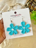 A closeup of a pair of bright sea blue crocheted, 5 petal flower-shaped dangle earrings by Chirpyknots, displayed on a white brand card over a woven mat and white fabric and pearl beads.