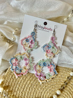 A pair of pastel multicoloured tatted, big size, heavily beaded dangle earrings by Chirpyknots, displayed on a white brand card over a woven mat and white fabric and pearl beads.