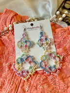 A closeup of a pair of pastel multicoloured tatted, big size, heavily beaded dangle earrings by Chirpyknots, displayed on a white brand card styled with peach color chikankari kurti