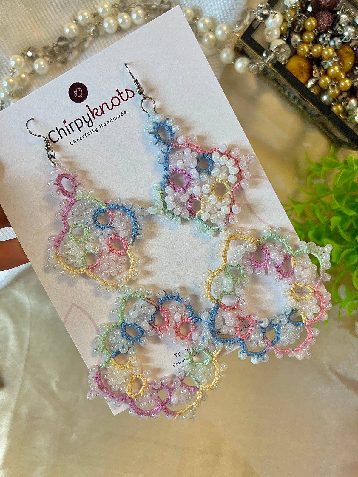 A closeup of a pair of pastel multicoloured tatted, big size, heavily beaded dangle earrings by Chirpyknots, displayed on a white brand card.