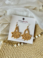 A pair of golden crocheted, dangle earrings by Chirpyknots, featuring a round floral shape with a delicate loop above it, displayed on a white brand card over a woven mat and white fabric and pearl beads.