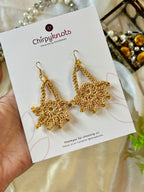 A closeup of a pair of golden crocheted, dangle earrings by Chirpyknots, featuring a round floral shape with a delicate loop above it, displayed on a white brand card.