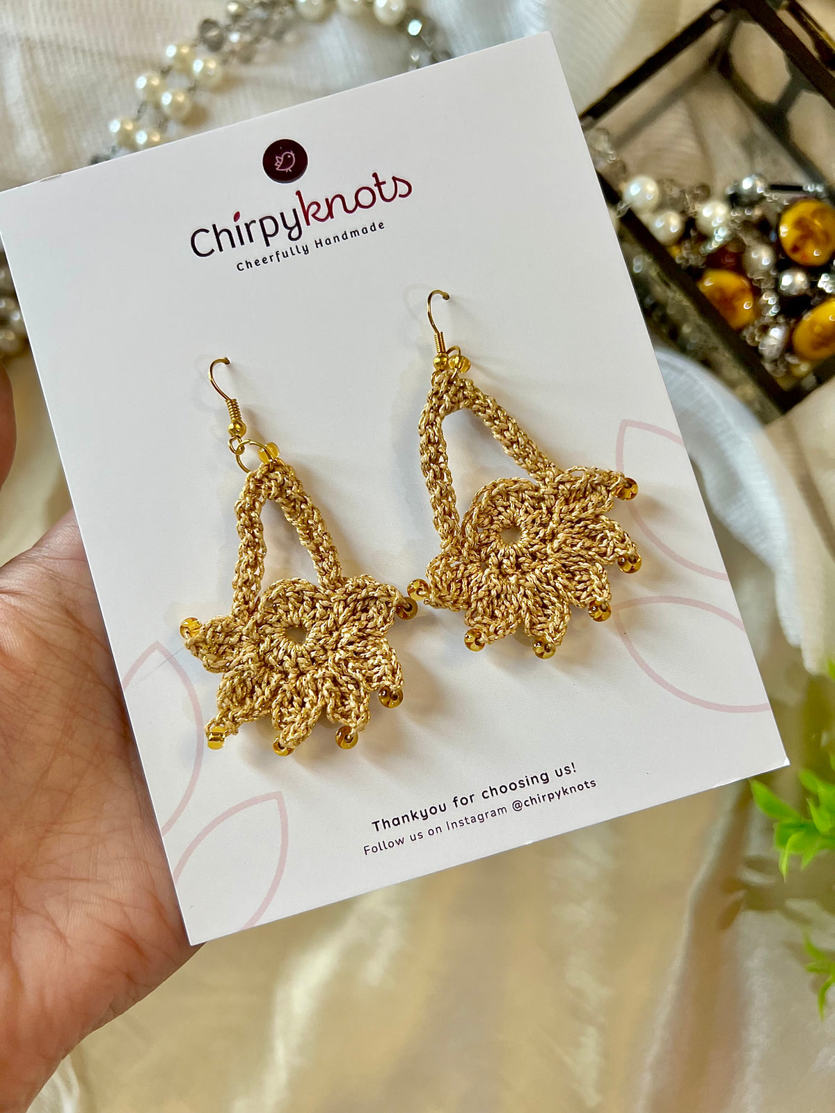A closeup of a pair of golden crocheted, dangle earrings by Chirpyknots, featuring a round floral shape with a delicate loop above it, displayed on a white brand card.