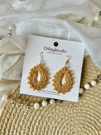 A pair of crocheted golden, oval shaped beaded dangle earrings by Chirpyknots, displayed on a white brand card over a woven mat and white fabric and pearl beads.