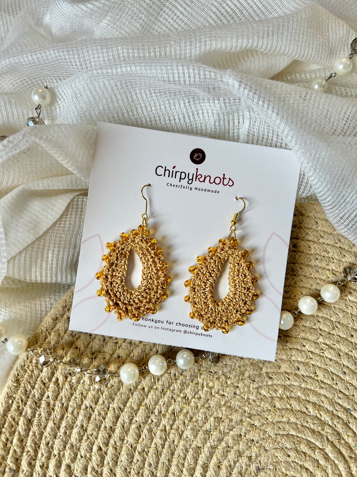 A pair of crocheted golden, oval shaped beaded dangle earrings by Chirpyknots, displayed on a white brand card over a woven mat and white fabric and pearl beads.