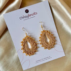 A closeup of a pair of crocheted golden, oval shaped beaded dangle earrings by Chirpyknots, displayed on a white brand card.