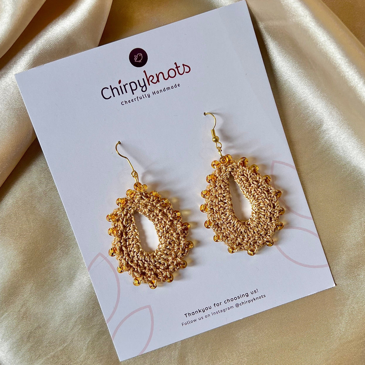 A closeup of a pair of crocheted golden, oval shaped beaded dangle earrings by Chirpyknots, displayed on a white brand card.