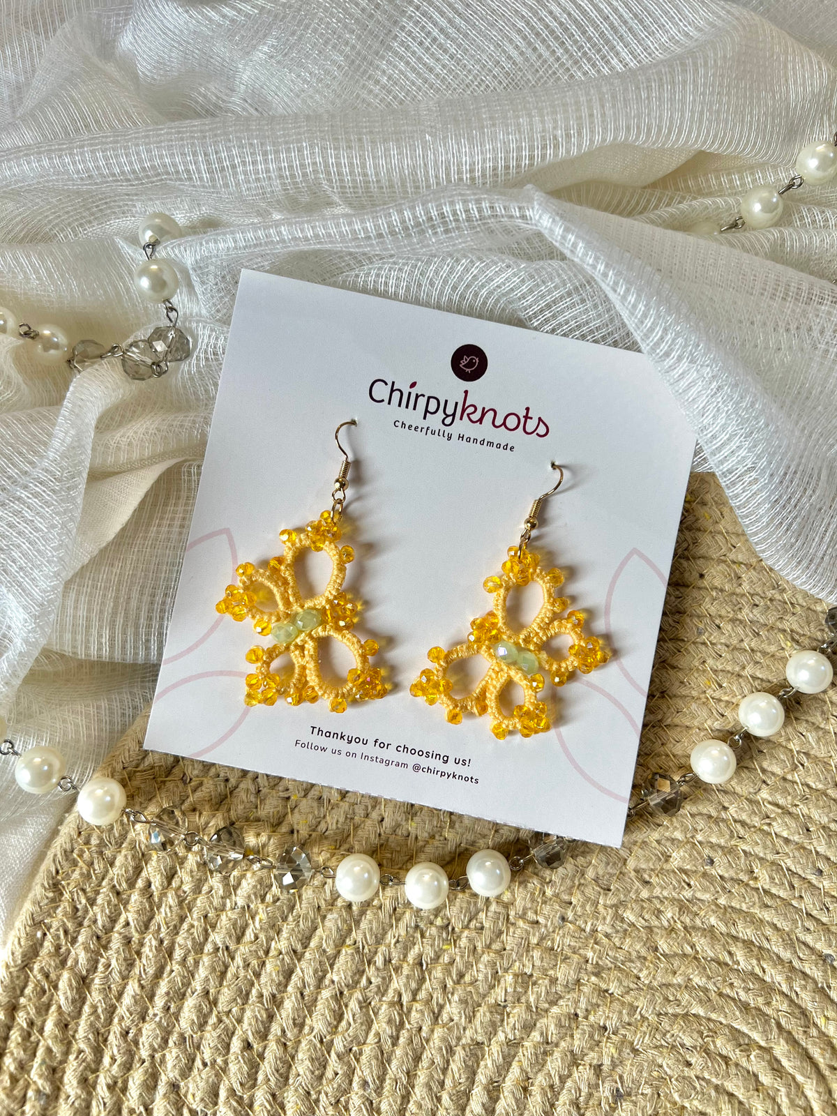 A pair of tatted pink, beaded, butterfly style dangle earrings by Chirpyknots, displayed on a white brand card over a woven mat and white fabric and pearl beads.