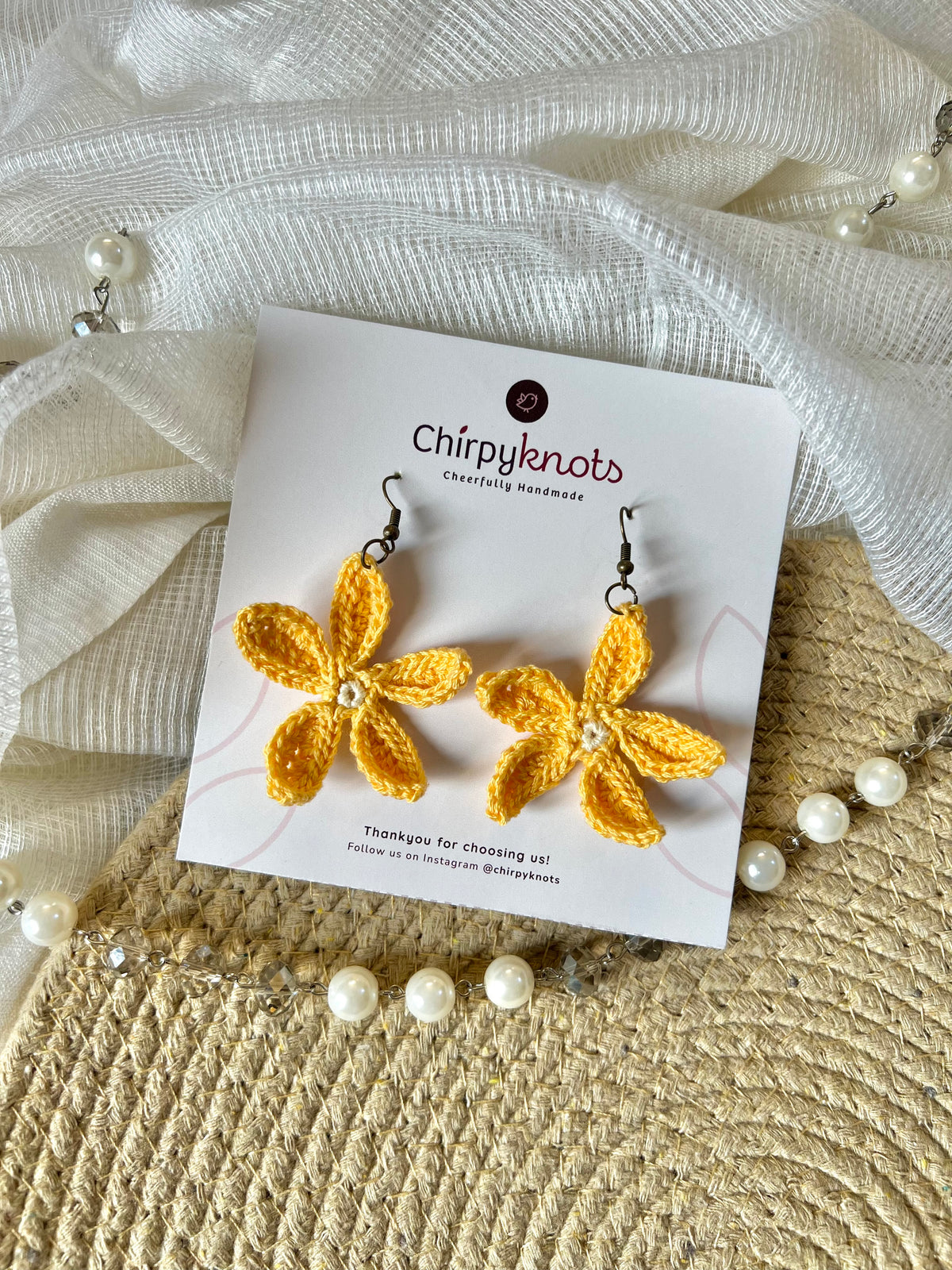 A pair of yellow crocheted, 5 petal flower-shaped dangle earrings by Chirpyknots, displayed on a white brand card over a woven mat and white fabric and pearl beads.
