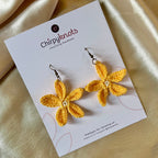 A closeup of a pair of yellow crocheted, 5 petal flower-shaped dangle earrings by Chirpyknots, displayed on a white brand card.