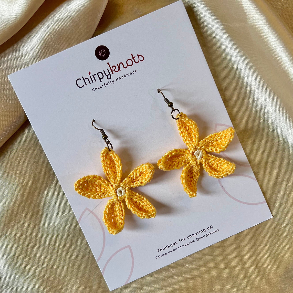 A closeup of a pair of yellow crocheted, 5 petal flower-shaped dangle earrings by Chirpyknots, displayed on a white brand card.
