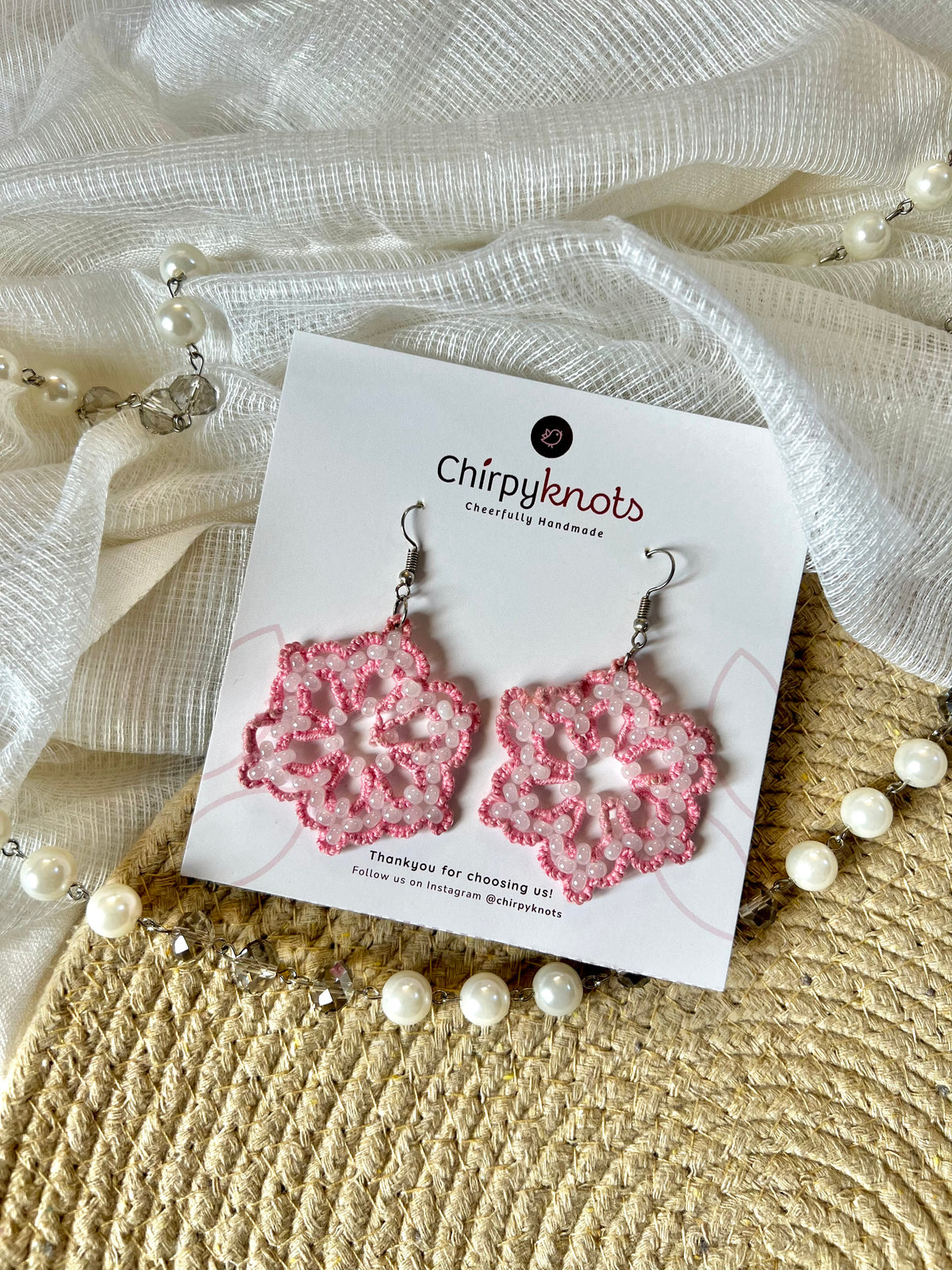 A pair of pink tatted, snowflake, beaded dangle earrings by Chirpyknots, displayed on a white brand card over a woven mat and white fabric and pearl beads.