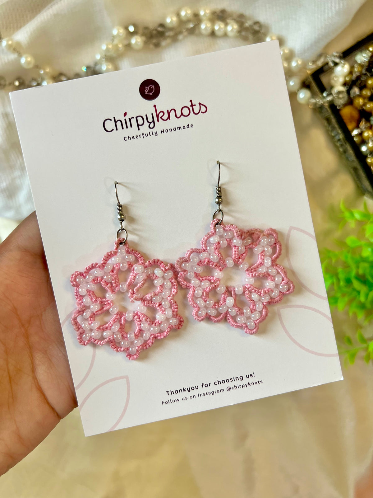 A closeup of a pair of pink tatted, snowflake, beaded dangle earrings by Chirpyknots, displayed on a white brand card.