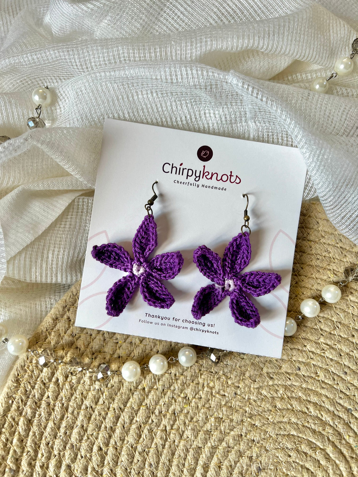 A pair of purple crocheted, 5 petal flower-shaped dangle earrings by Chirpyknots, displayed on a white brand card over a woven mat and white fabric and pearl beads.