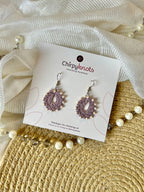 A pair of crocheted lavender, small, oval shaped beaded dangle earrings by Chirpyknots, displayed on a white brand card over a woven mat and white fabric and pearl beads.
