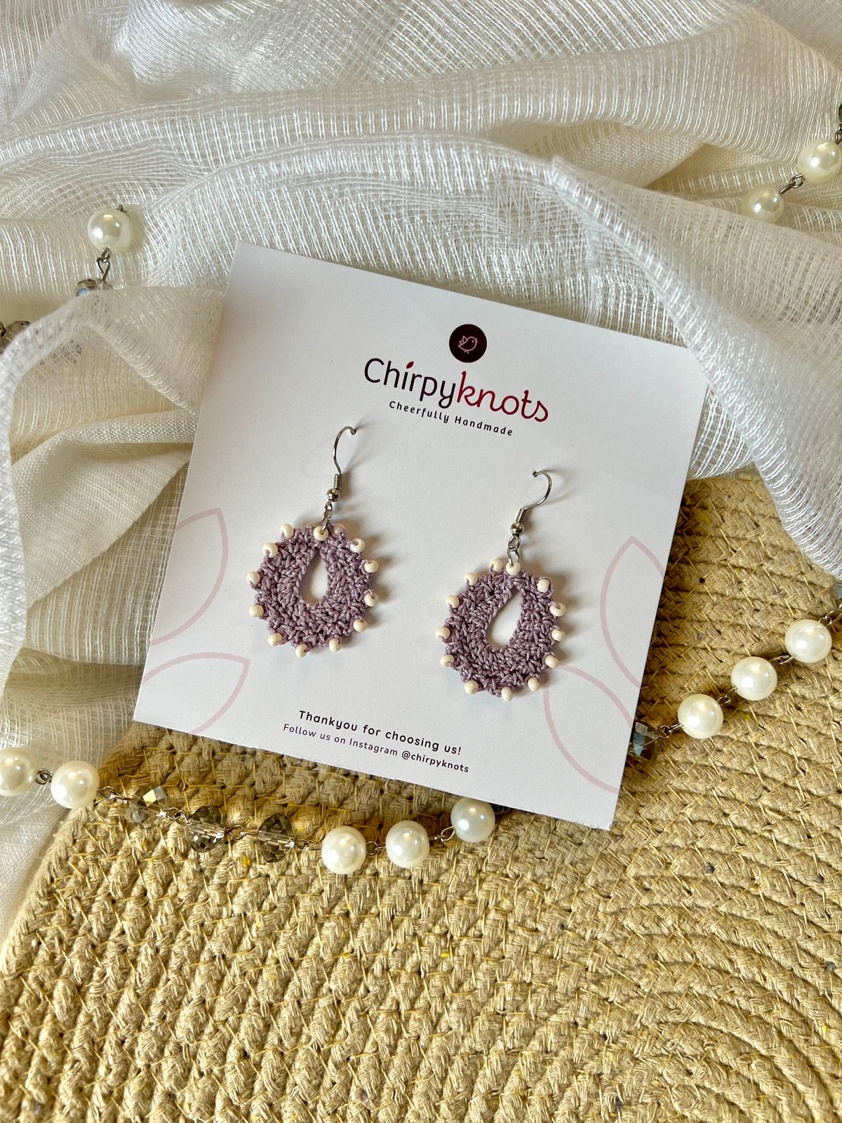 A pair of crocheted lavender, small, oval shaped beaded dangle earrings by Chirpyknots, displayed on a white brand card over a woven mat and white fabric and pearl beads.